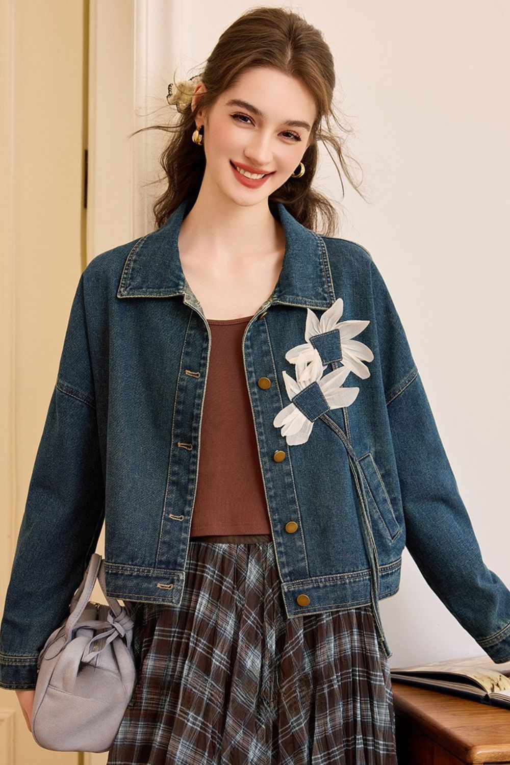 Woman wearing a denim jacket with large white floral appliqué, brown top, and plaid skirt, smiling and carrying a gray handbag