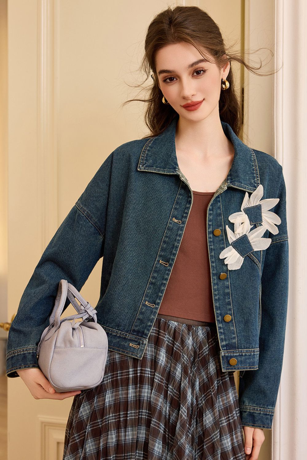 Model standing in a denim jacket with floral appliqué, brown top, and plaid skirt, holding a gray handbag