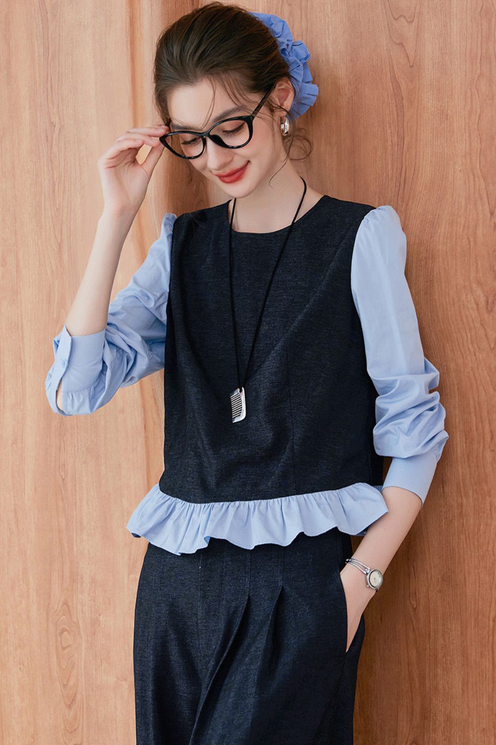 Model adjusting glasses while wearing navy blue vest and pants with light blue ruffle-trim blouse