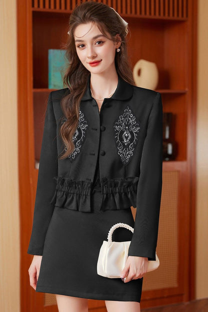 Indoor pose of model in black embroidered jacket with tassels and black skirt
