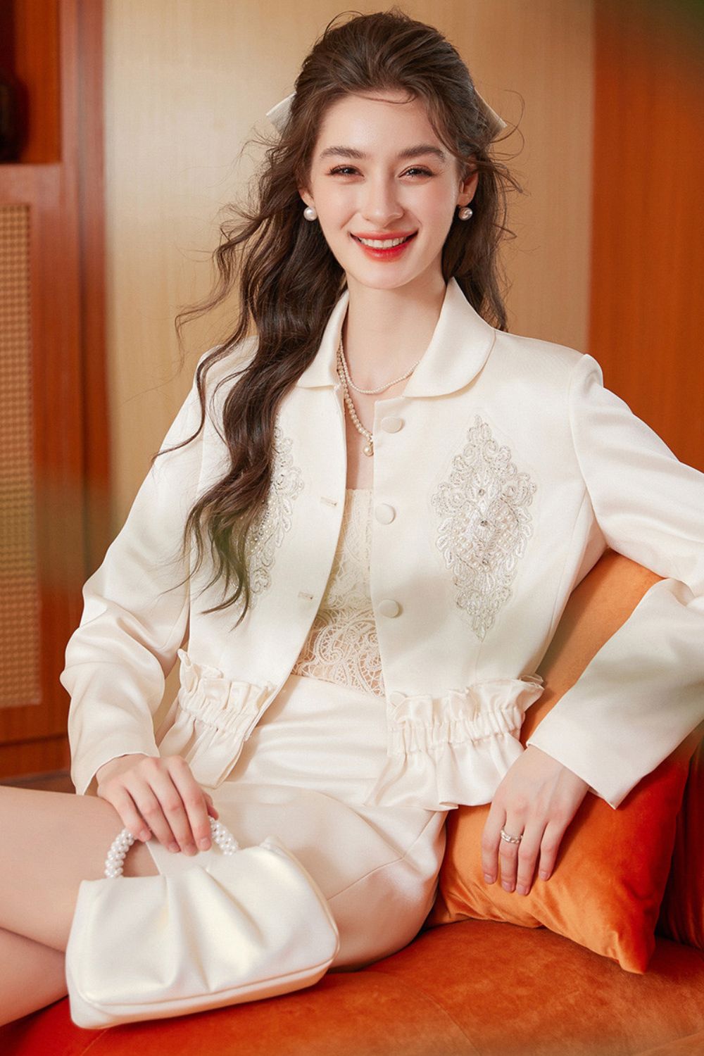 Smiling model in cream embroidered tassel jacket and cream mini skirt seated on a sofa