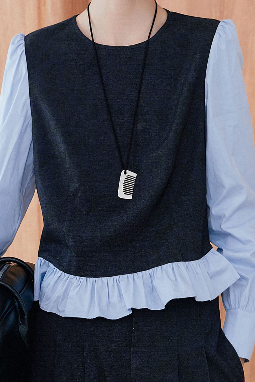 Close-up of navy blue sleeveless vest over light blue blouse with ruffled hem, accessorized with a pendant necklace